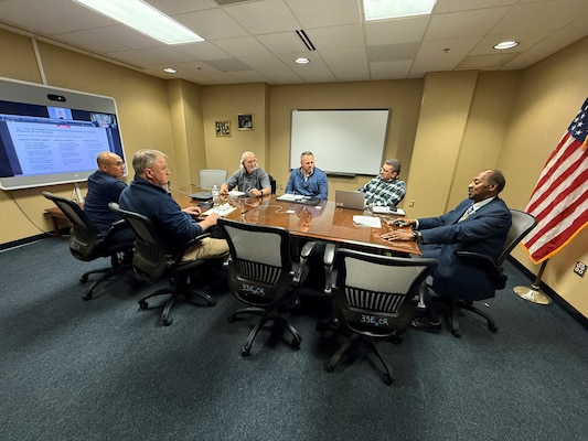 A group of seated people watch a teleconference in a conference room.