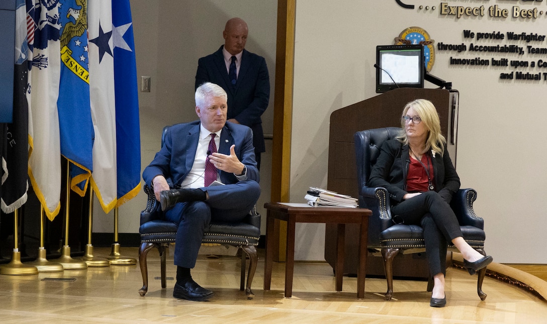 A man and a woman sit in chairs on a stage.