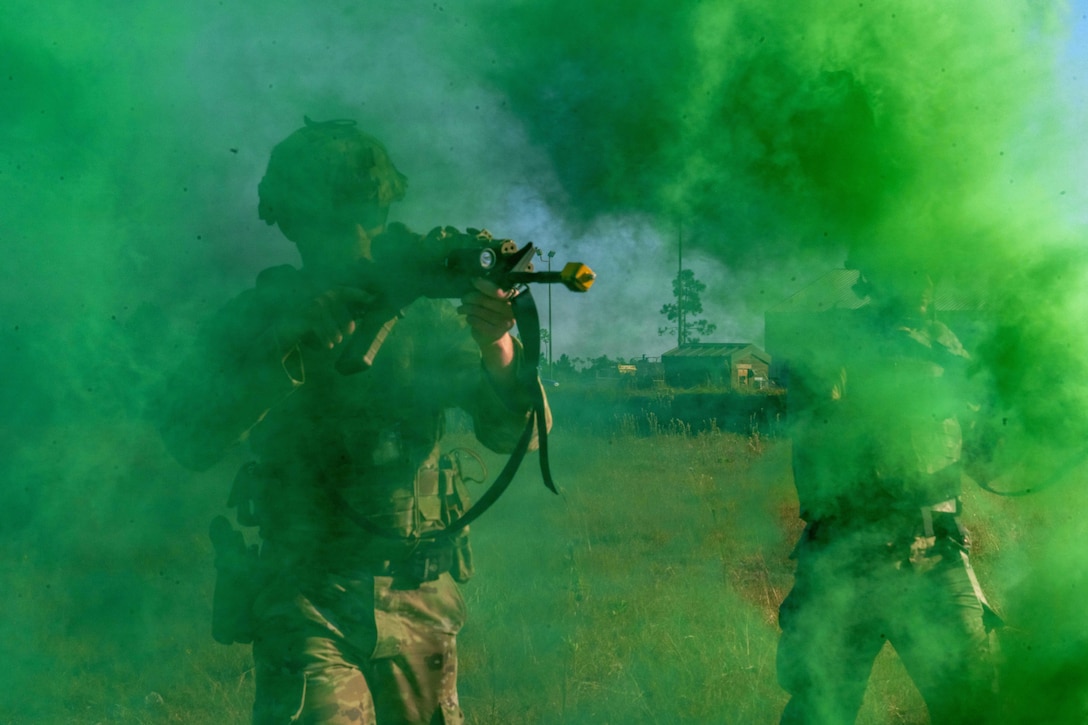 Two airmen in camouflage combat gear and aiming rifles walk through green plumes of smoke outdoors during daytime, with a building in the background.