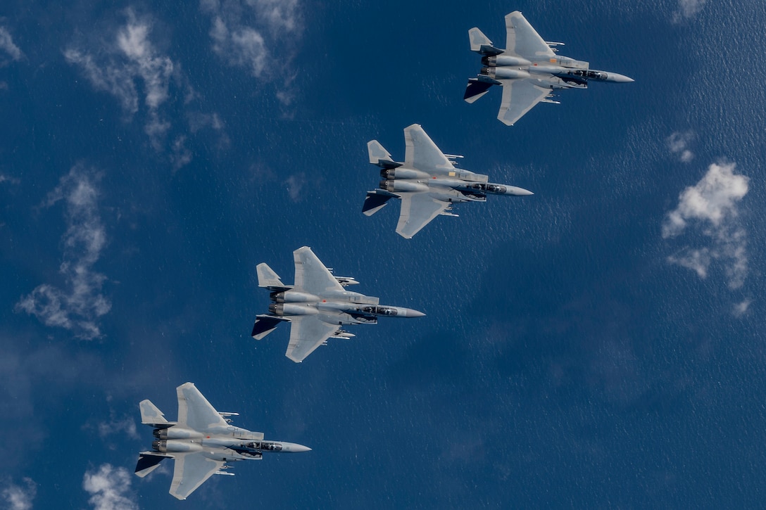 Four jets fly in formation over a blue body of water, with white clouds visible in the sky below.