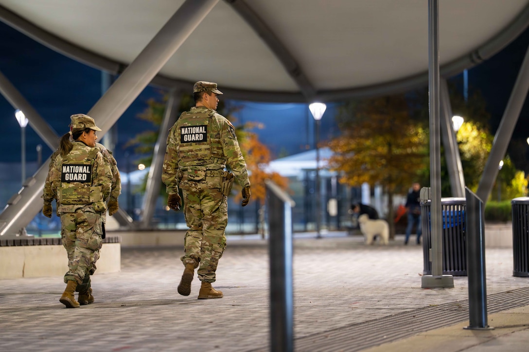Three guardsmen walk along a covered paved area in dark conditions, with trash cans, lit lampposts, small trees, other people and a dog in the background.