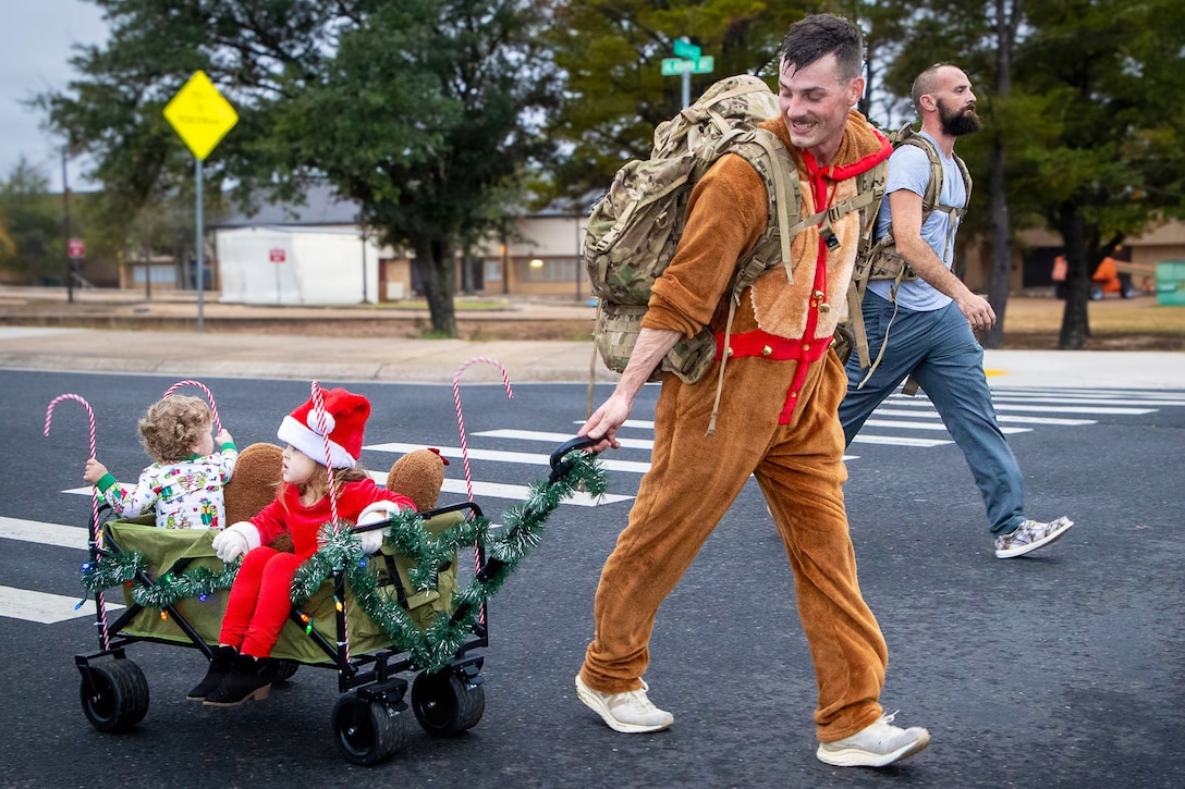 A person wearing a brown and tan costume and backpack pulls a wagon with two children sitting in it while walking on a street during daytime, with another person in civilian clothing and also carrying a backpack walking nearby.