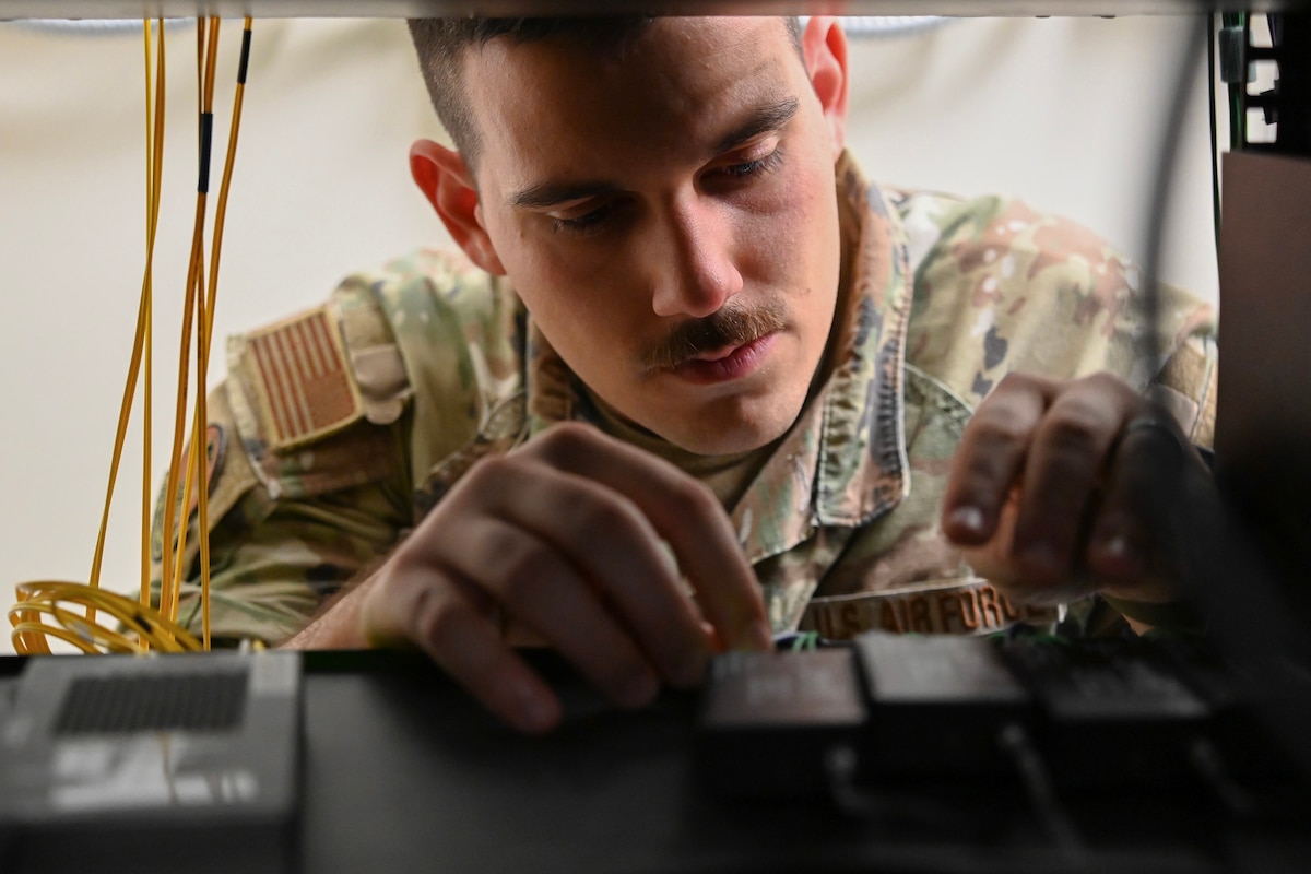 A uniformed service member reaches his hands and peers into a shelf with server connection sources.