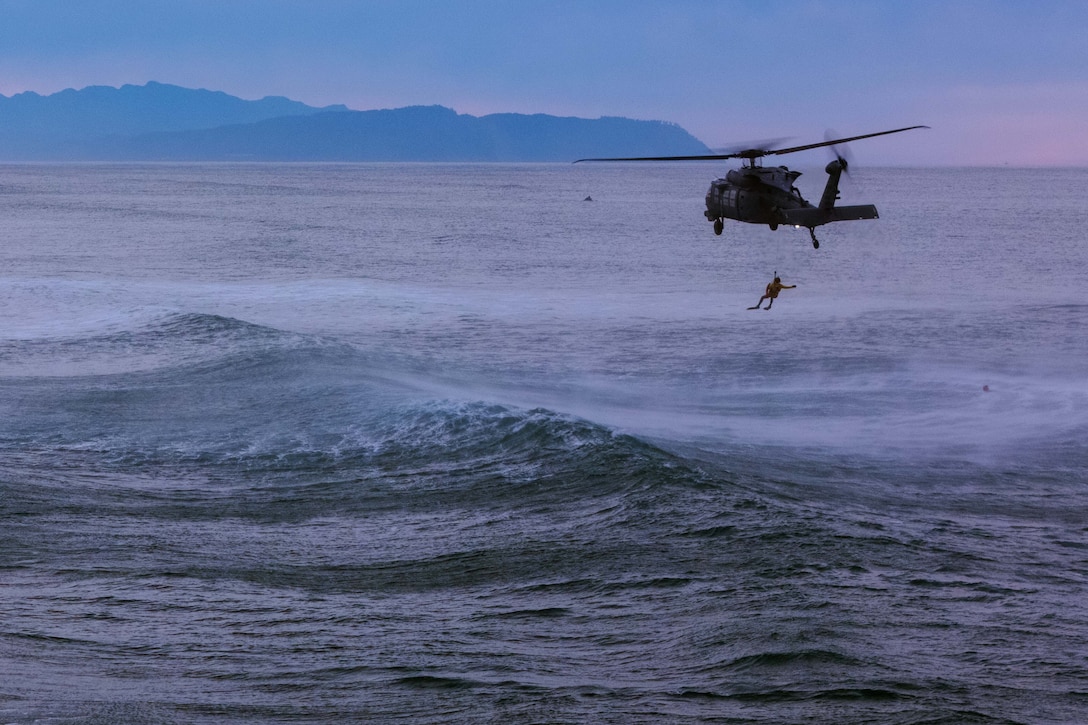 A person attached to a cable dangles from a flying helicopter over a wavy body of water, with a hazy blue and pink sky and hilly land in the background.