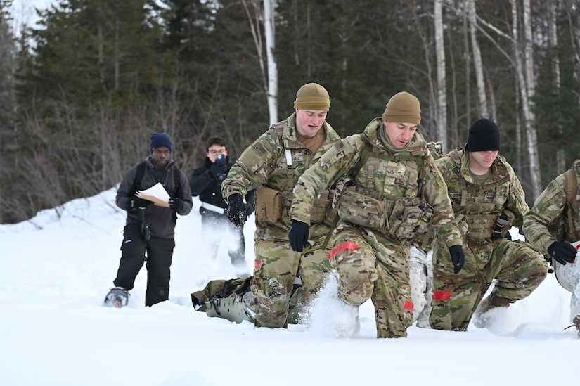 Four people in combat gear haul a litter in knee-deep snow. Two people behind them monitor their progress and film them.