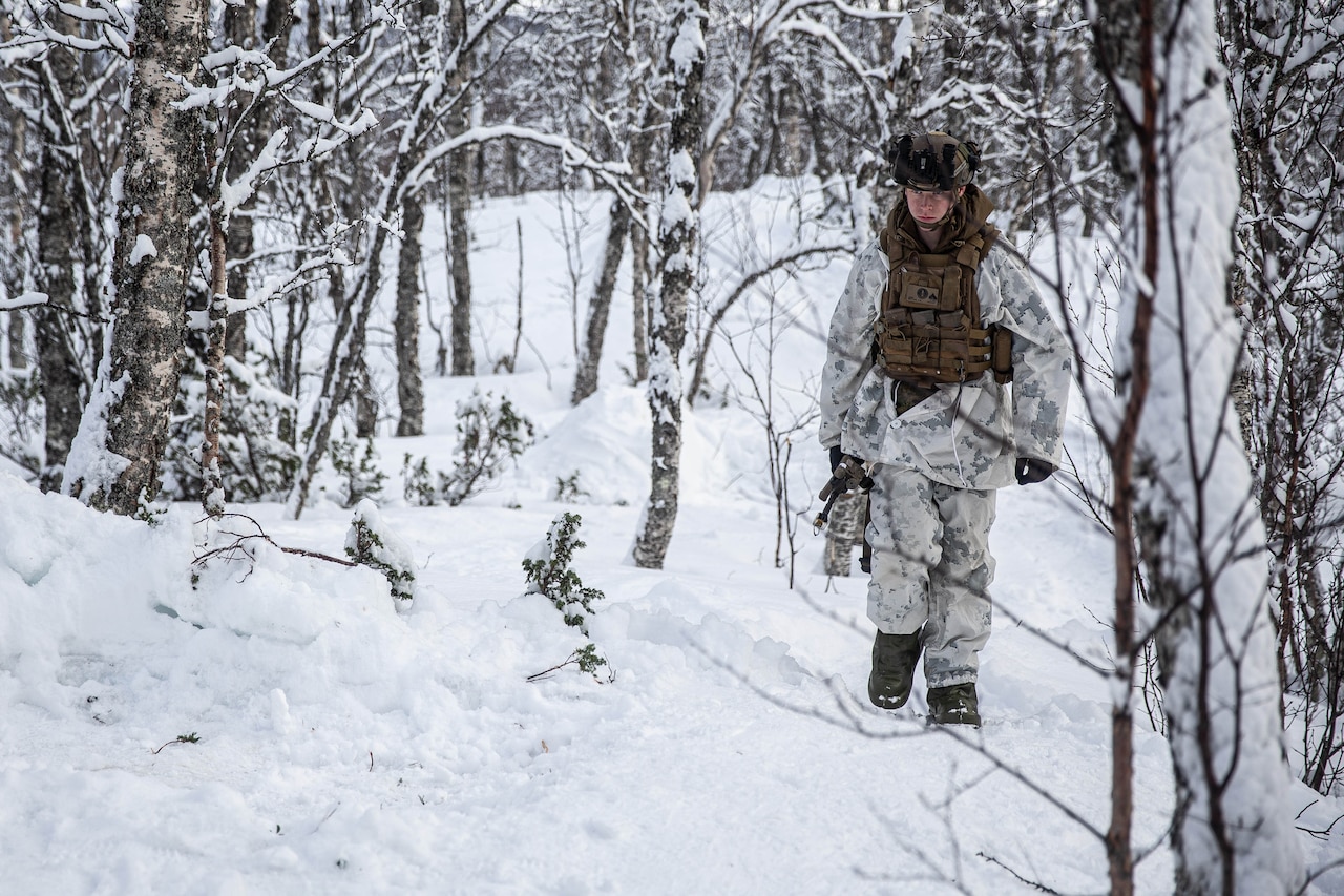 A man in heavy-duty winter combat gear walks through a snow-covered forest.