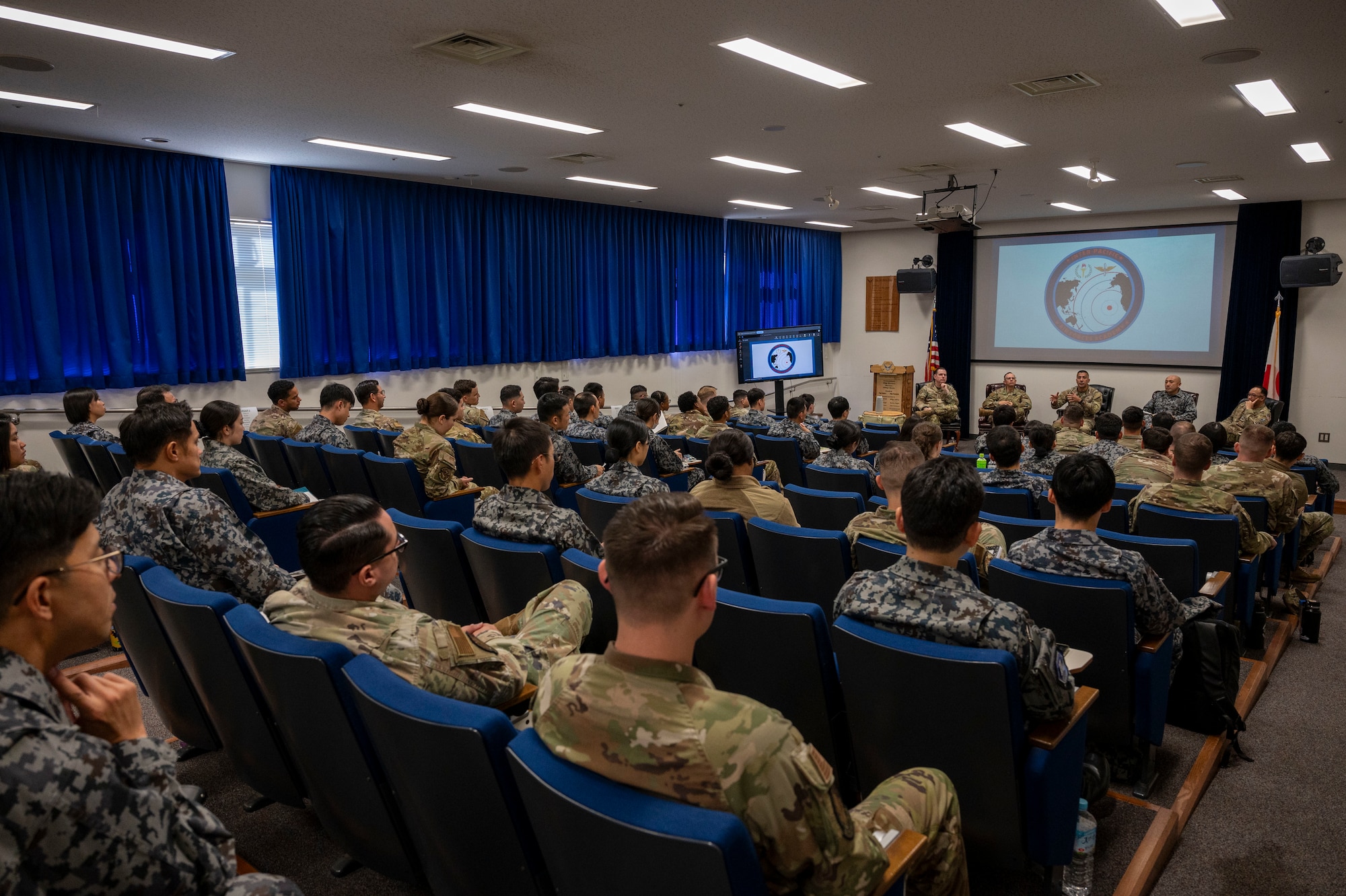 A group of military members sit in a room as five military members in the front of the room speak.