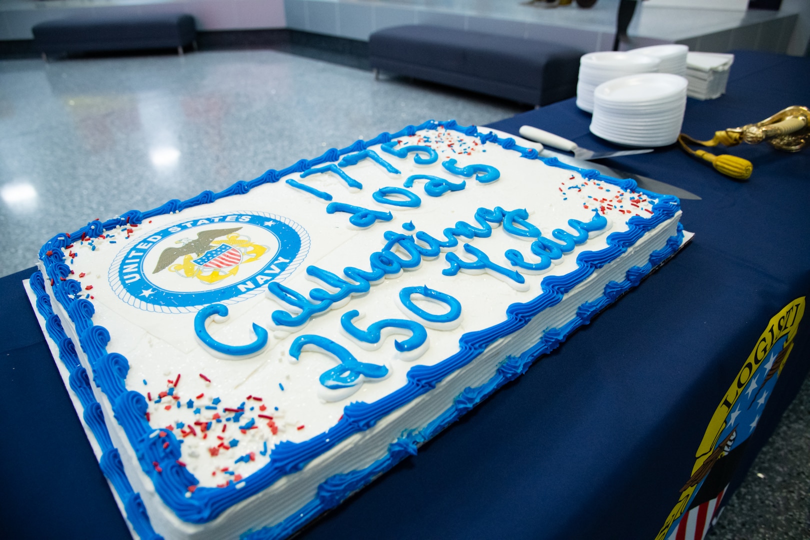 A white sheet cake that reads "1775-2025 Celebrating 250 Years" with the U.S. Navy seal on it sits on a table with a blue tablecloth.