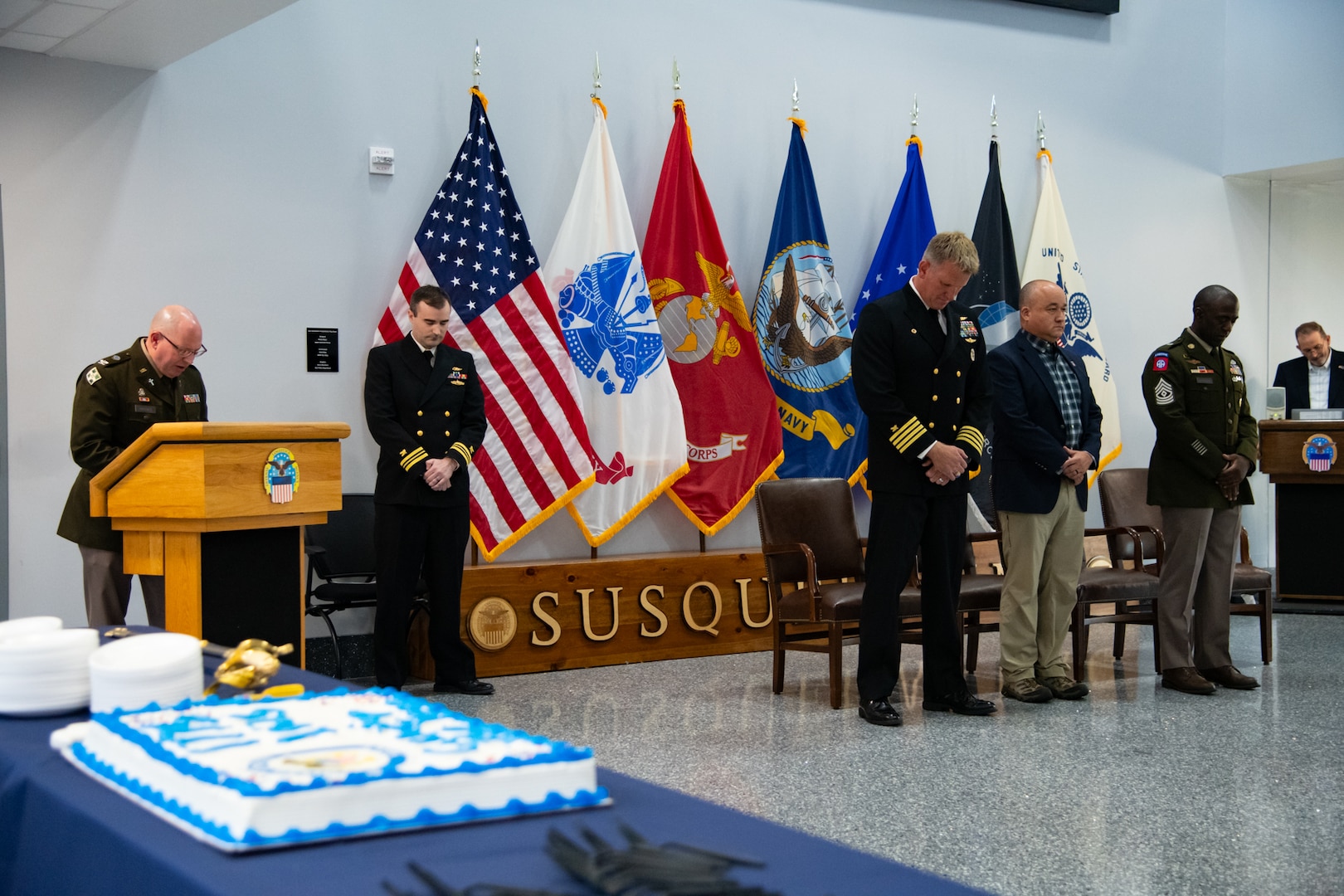 A man wearing a U.S. Army Green Service Uniform stands behind a lectern speaking to an audience.