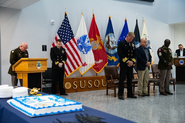 A man wearing a U.S. Army Green Service Uniform stands behind a lectern speaking to an audience.