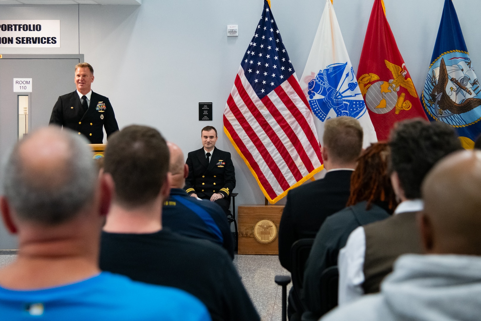 A man wearing a U.S. Navy Service Dress Blue uniform stands behind a lectern speaking to an audience.