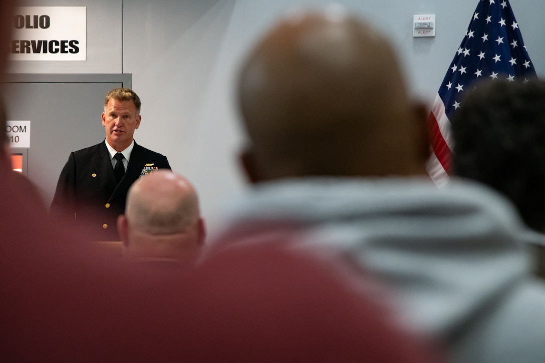 A man wearing a U.S. Navy Service Dress Blue uniform stands behind a lectern speaking to an audience.