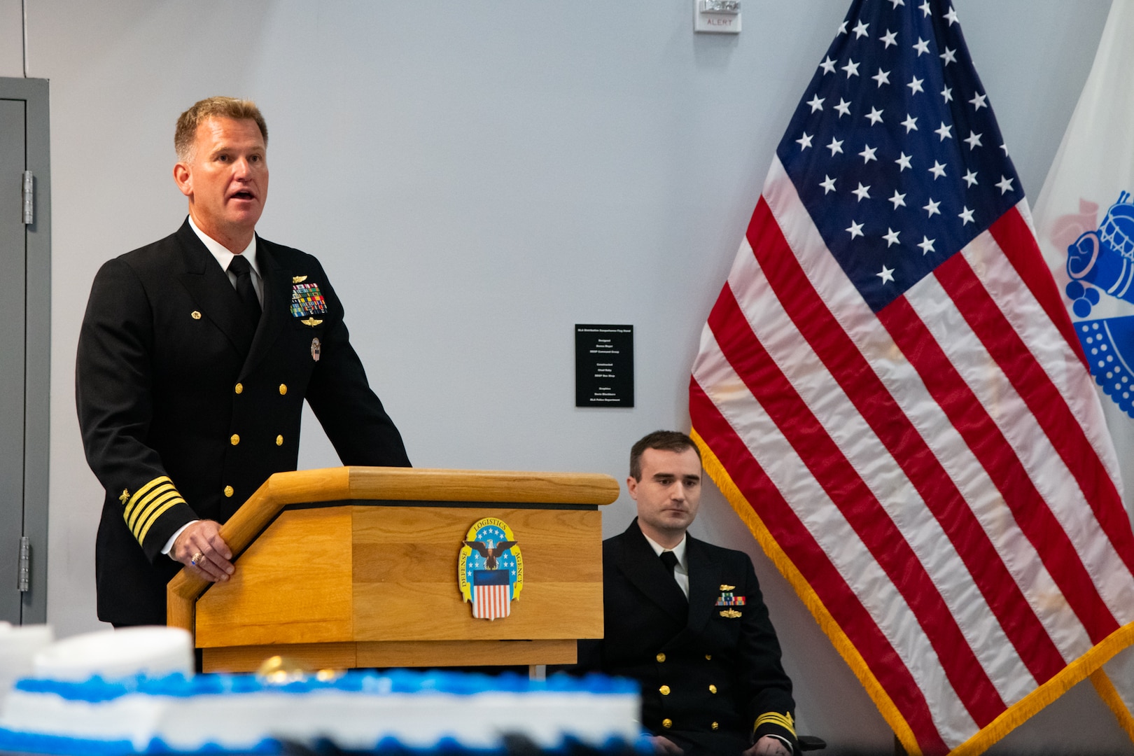 A man wearing a U.S. Navy Service Dress Blue uniform stands behind a lectern speaking.