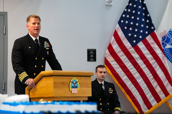 A man wearing a U.S. Navy Service Dress Blue uniform stands behind a lectern speaking.