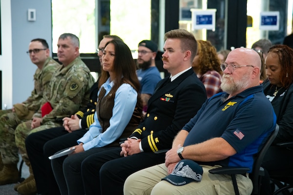 A group of people seated indoors in rows look toward a person speaking.
