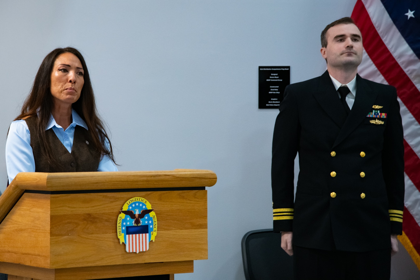 A woman stands behind a lectern speaking. There is a man wearing a U.S. Navy Service Dress Blue uniform to her left.