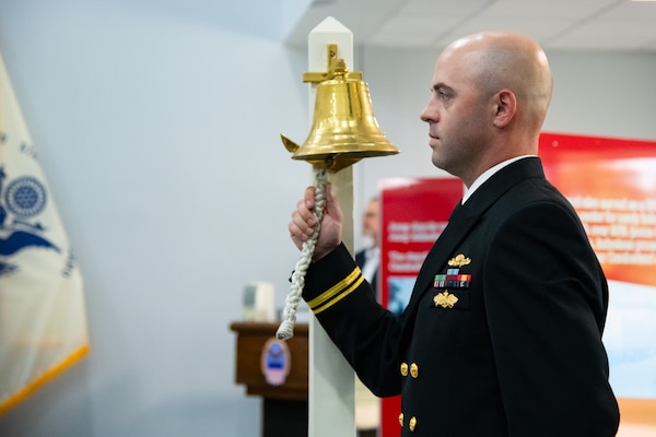 A man wearing a U.S. Navy Service Dress Blue uniform holds the bell rope of a brass bell.
