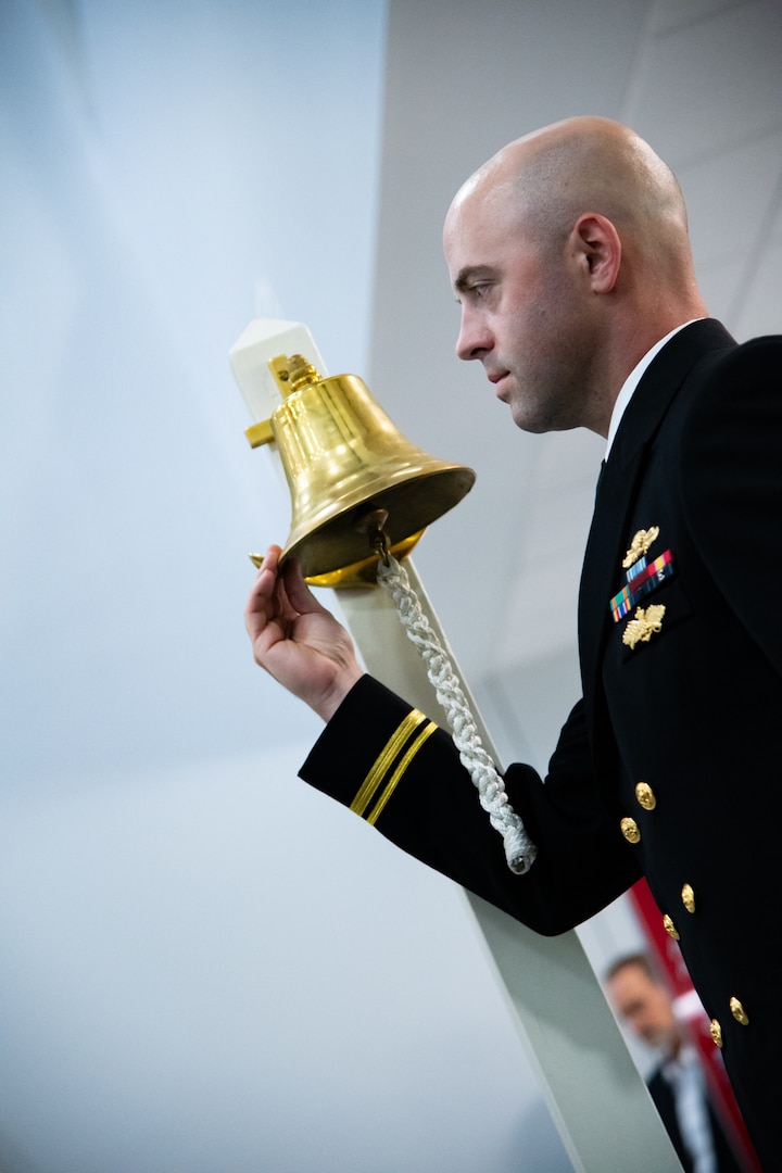 A man wearing a U.S. Navy Service Dress Blue uniform holds the lip of a brass bell.