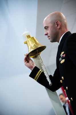 A man wearing a U.S. Navy Service Dress Blue uniform holds the lip of a brass bell.