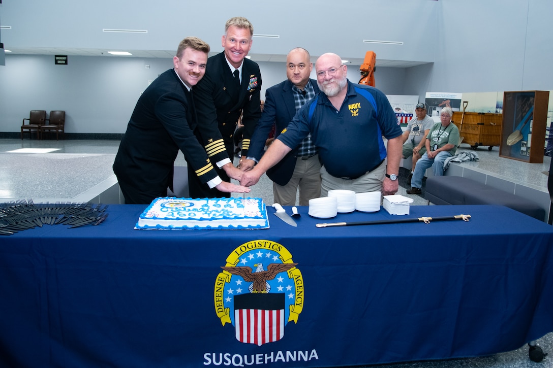 Four men, two wearing military uniforms, stand behind a table with a blue tablecloth cutting a cake that's on the table.