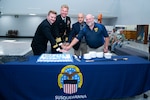 Four men, two wearing military uniforms, stand behind a table with a blue tablecloth cutting a cake that's on the table.
