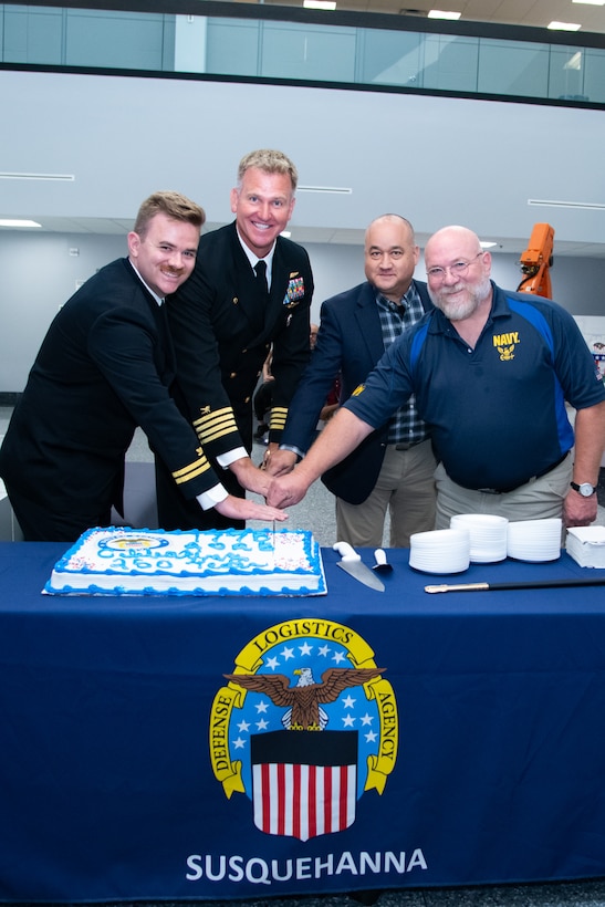 Four men, two wearing military uniforms, stand behind a table with a blue tablecloth cutting a cake that's on the table.