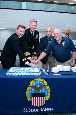 Four men, two wearing military uniforms, stand behind a table with a blue tablecloth cutting a cake that's on the table.