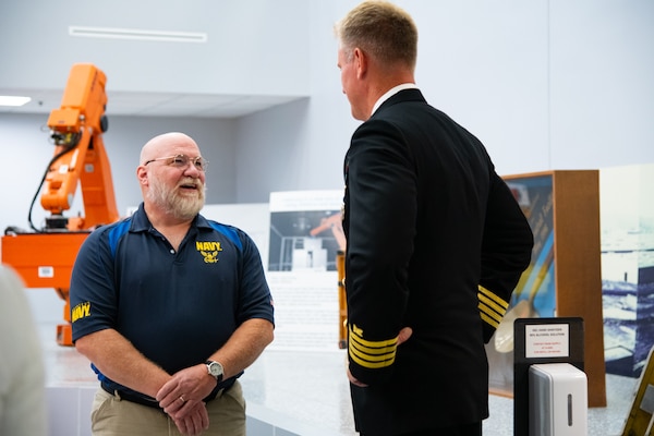 Two men stand indoors speaking. One is wearing a U.S. Navy Service Dress Blue uniform.