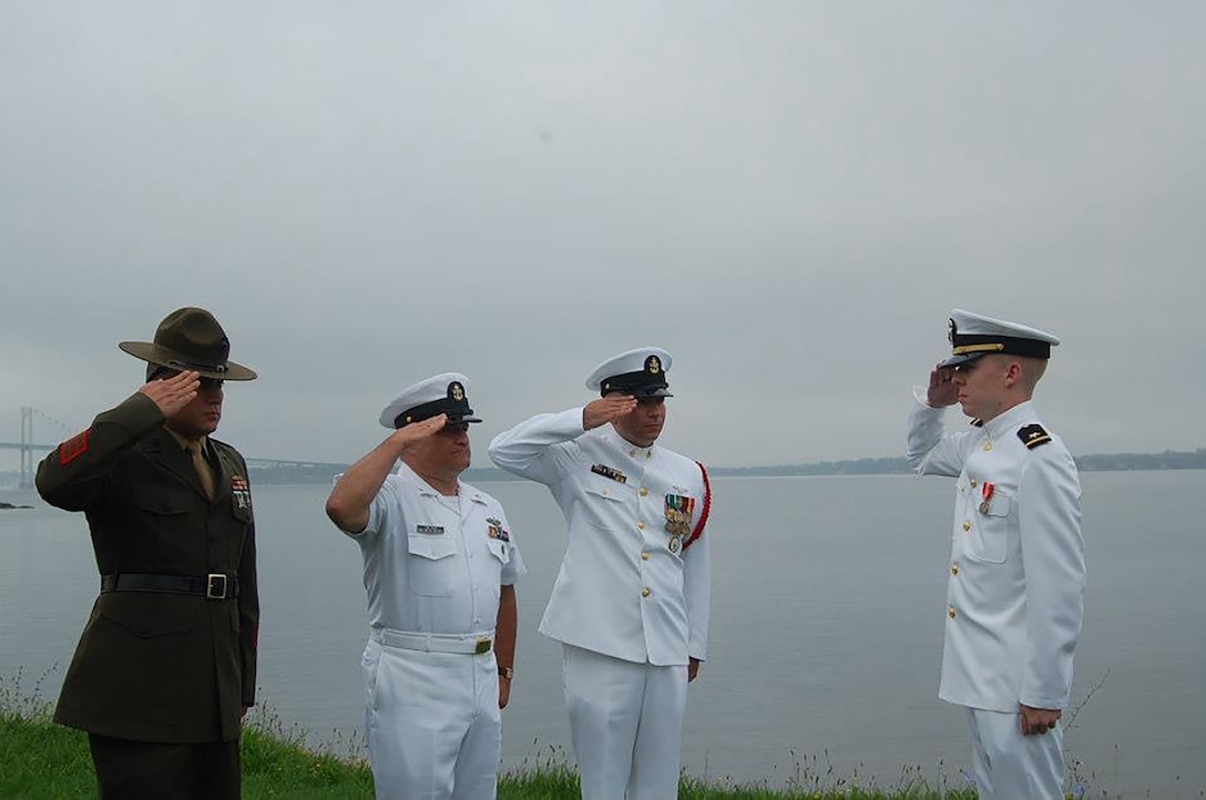 Four men wearing U.S. military uniforms stand outside on the shoreline saluting each other.
