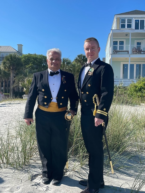 Two men in U.S. Navy uniforms stand outside on the beach.