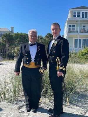 Two men in U.S. Navy uniforms stand outside on the beach.