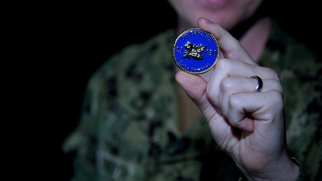 A man wearing a camouflage military uniform holds up a blue and gold coin.