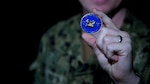 A man wearing a camouflage military uniform holds up a blue and gold coin.
