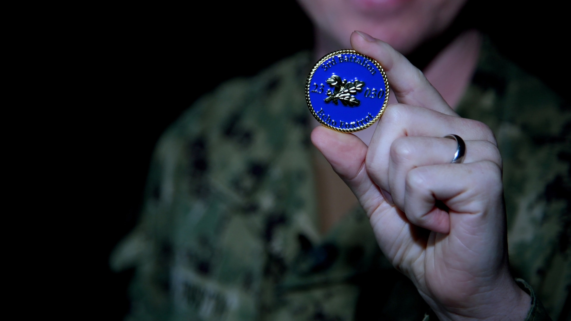 A man wearing a camouflage military uniform holds up a blue and gold coin.