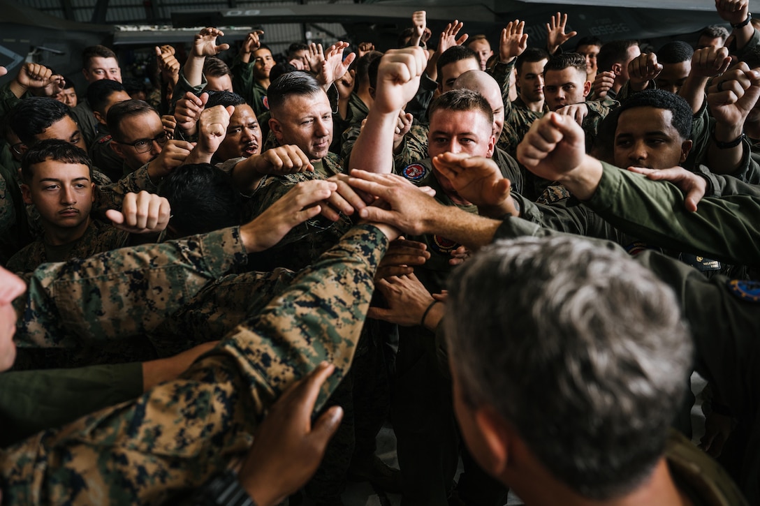 Marines raise their arms out towards the center of their group and touch hands inside a dimly lit building.