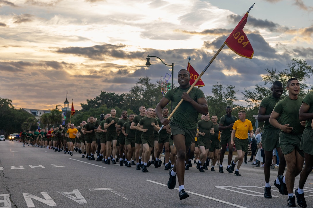 Marines in green t-shirts and shorts run on a street on a partly cloudy day while three hold red flags.