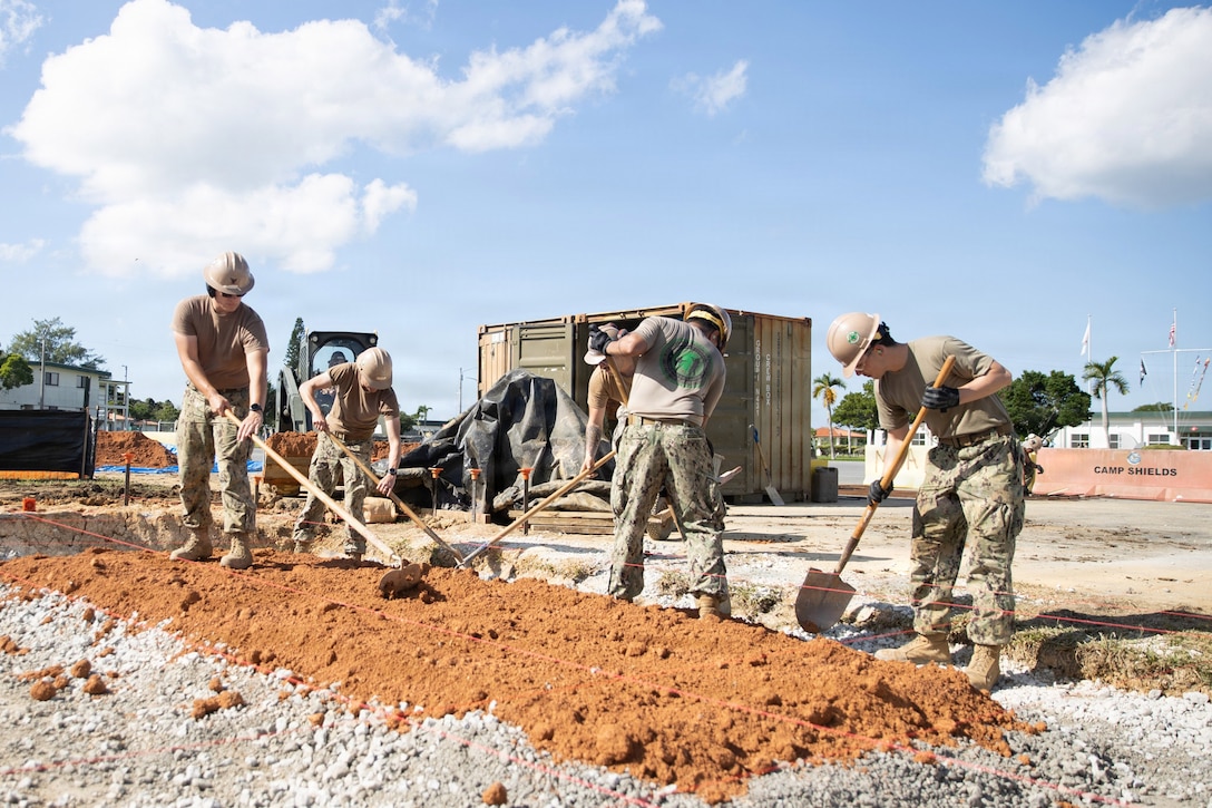 Five sailors use shovels to pack dirt onto gravel with a container and a construction vehicle behind on a sunny day.