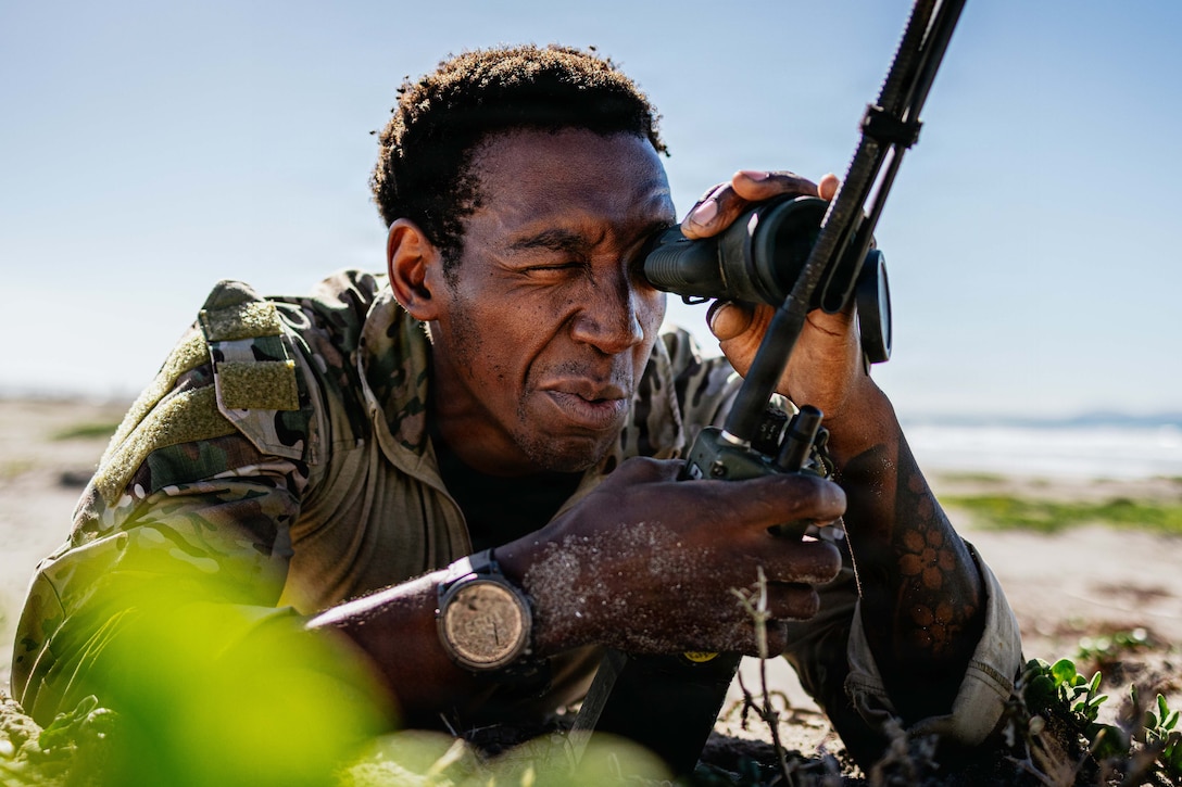 A sailor looks through a monocular device while leaning on their elbows near vegetation on a beach under a sunny sky.