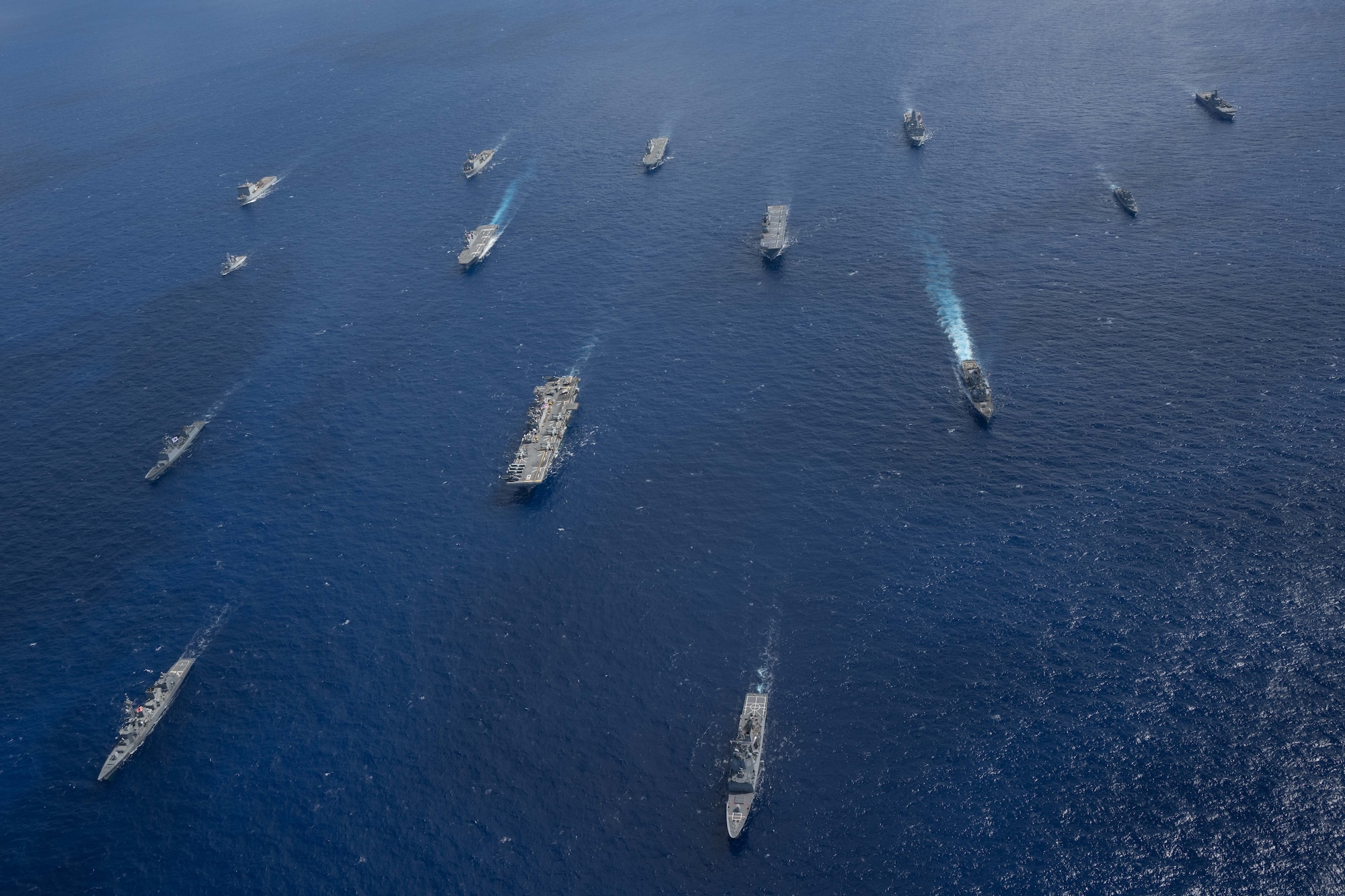 Over a dozen ships of various sizes cut through the ocean from an overhead view