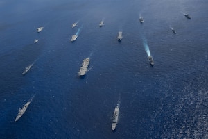 Over a dozen ships of various sizes cut through the ocean from an overhead view