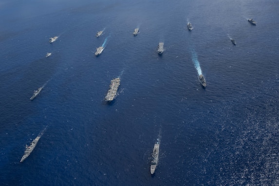 Over a dozen ships of various sizes cut through the ocean from an overhead view