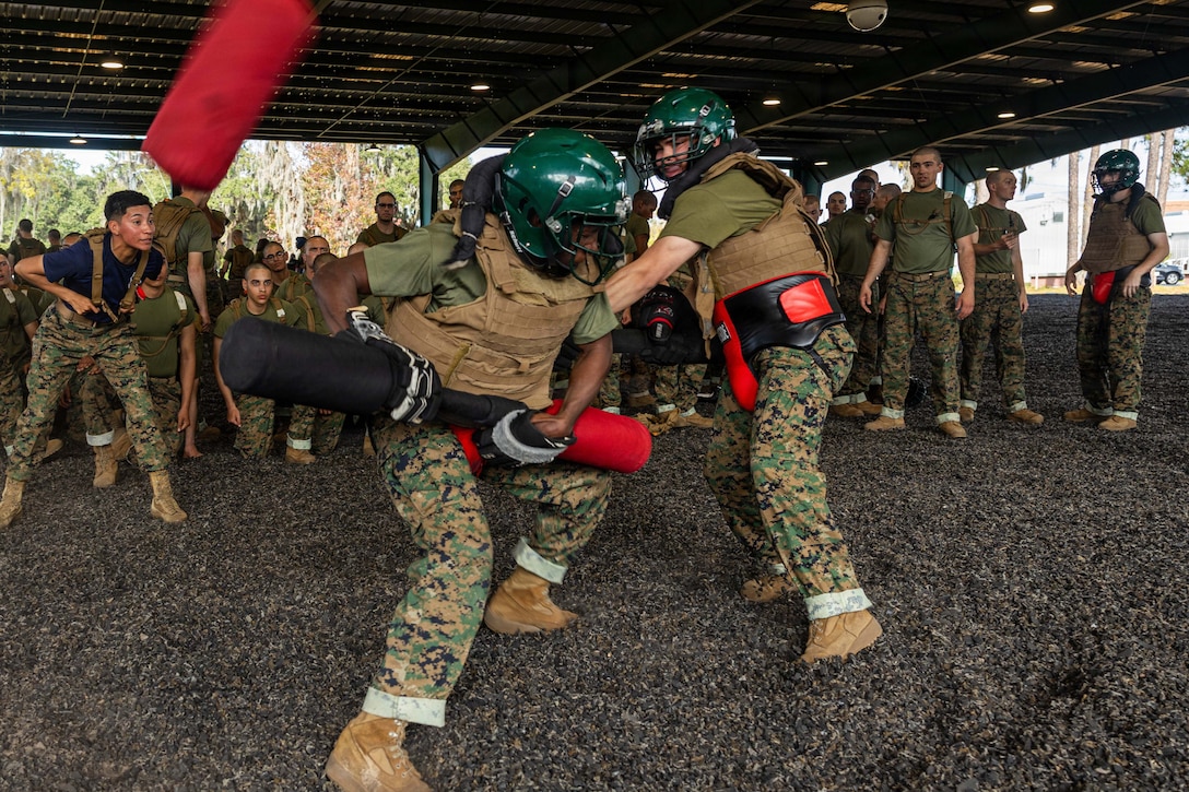 Two Marine recruits wearing head and body protection spar with pugil sticks as other Marine recruits stand or kneel and watch in a dirt area under a large awning outdoors.