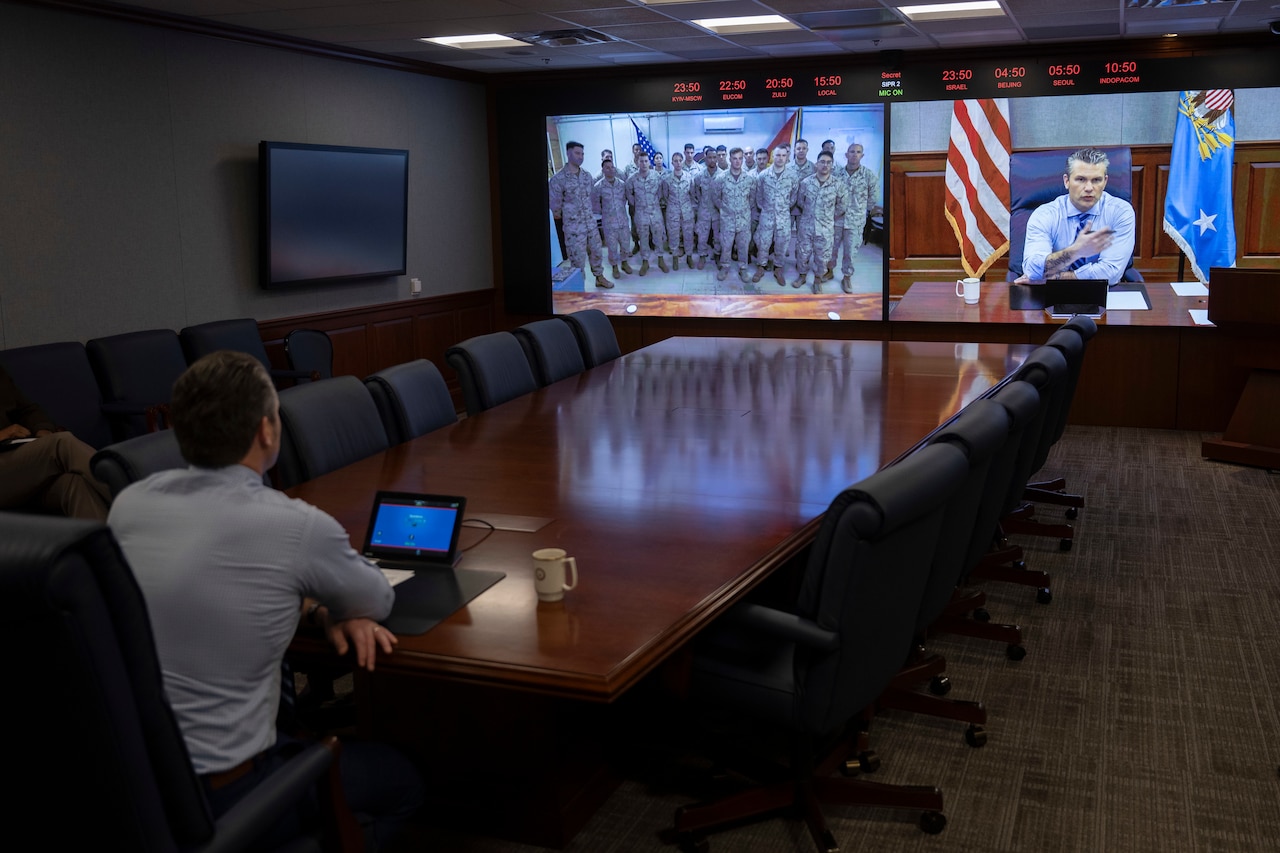 A man in business attire sitting at a conference table watches a large monitor that shows service members sitting in an auditorium. On the adjacent screen is the live video of the man sitting at the conference table.