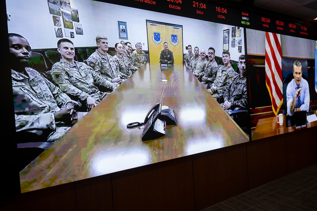 A large video monitor shows an image of service members sitting at a conference table. On the adjacent screen is a video of a man in business attire sitting at a conference table with a laptop on it.