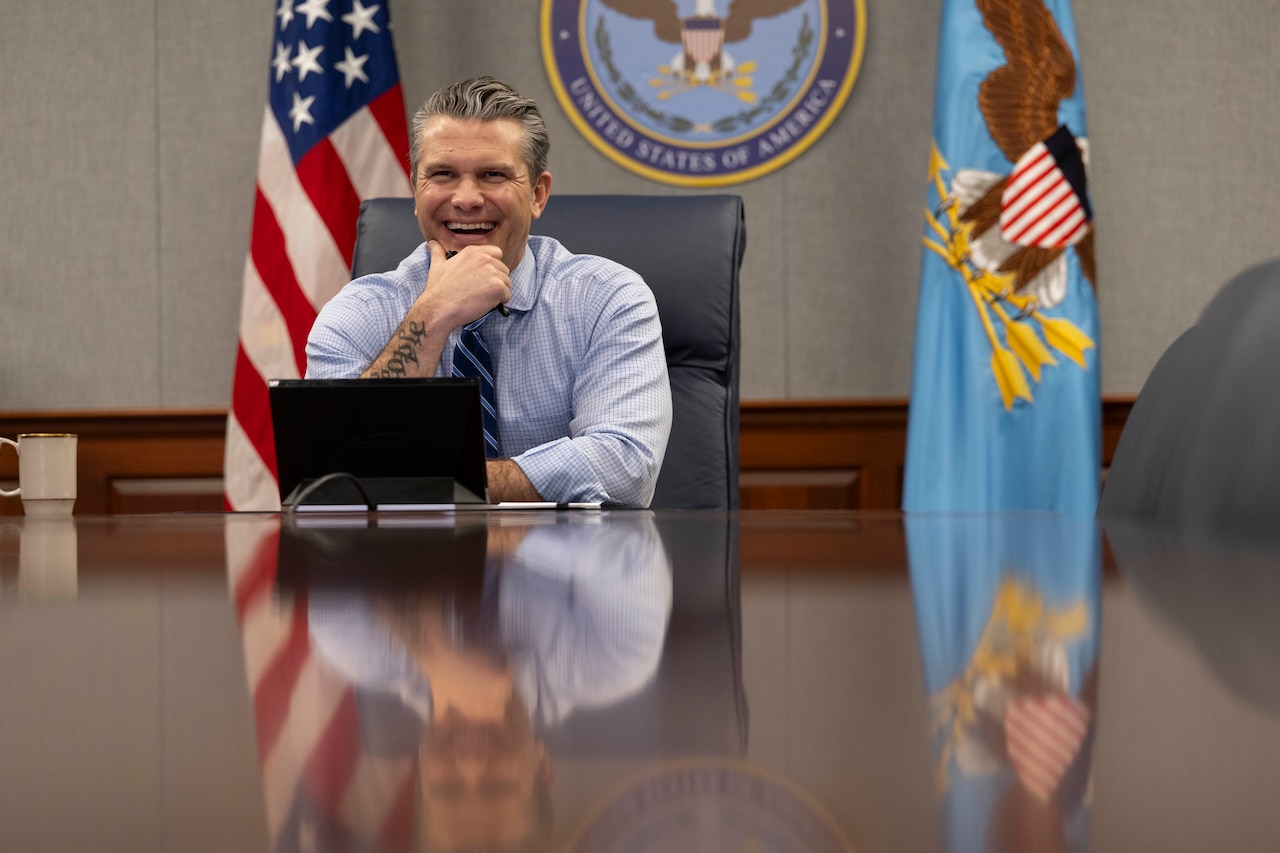 A smiling man in business attire sits at a conference table with a laptop on it.