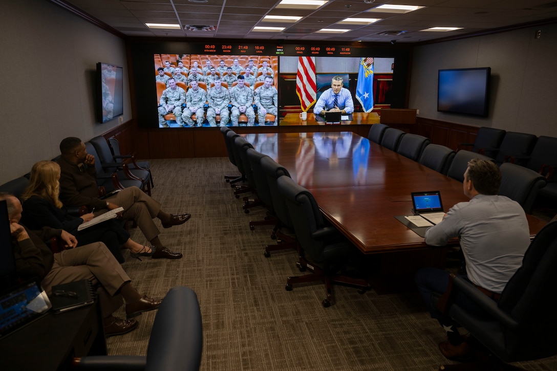 A man sitting at a conference table watches a large monitor that shows service members sitting in an auditorium. On the adjacent screen is a civilian man sitting at a conference table with a laptop on it.