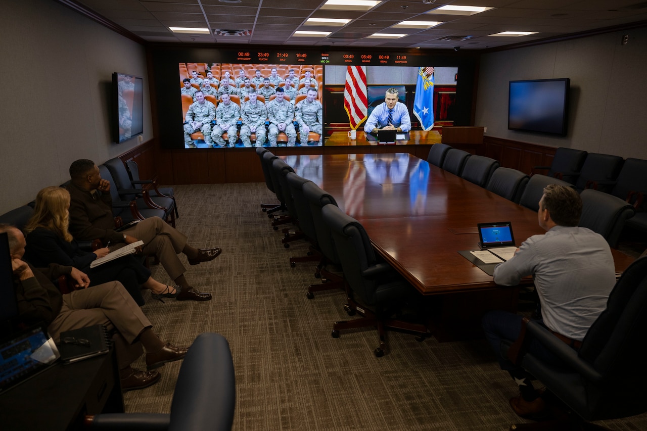 A man sitting at a conference table watches a large monitor that shows service members sitting in an auditorium. On the adjacent screen is a civilian man sitting at a conference table with a laptop on it.