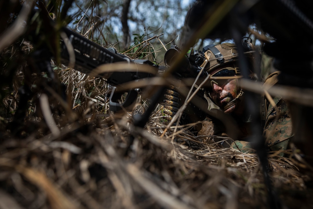 A Marine lying in a wooded area looks through the scope of a large machine gun during daytime.