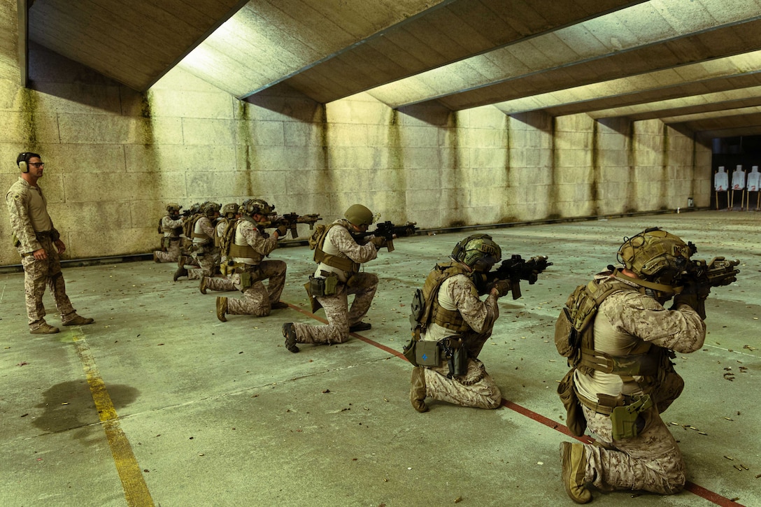 Kneeling Marines in combat gear fire rifles at targets in an enclosed concrete building, as a person wearing camouflage attire and ear protection stands behind them.