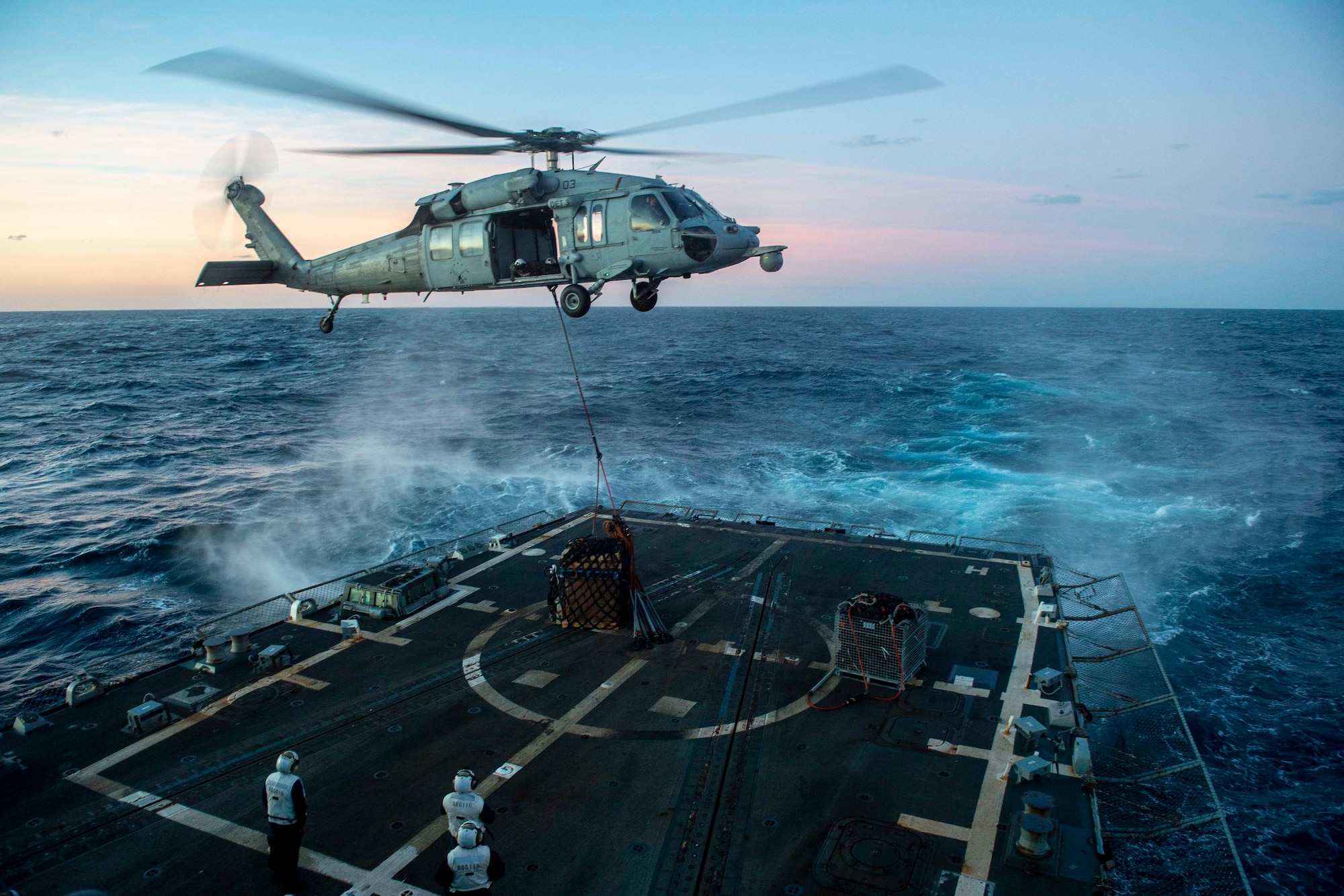 A helicopter stis up the waves around a boat at sunset as it drops off a large container suspended by a line